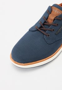 Chaussure textile bleu marine avec une surface texturée, accents en cuir marron, lacets marron et une semelle en caoutchouc blanche avec un contour beige.