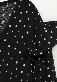 Tissu noir avec des pois blancs, présentant une surface texturée, un col en V et un détail d'épaule froncé.