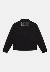 Svart puffjacka med hög krage, med horisontell sömnad och en stor vit Lacoste-logotyp på övre rygg. Elastiska ärmslut.
