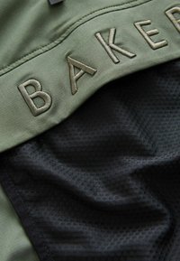 Tessuto verde oliva con "BAKER" ricamato in beige sopra una rete nera testurizzata. I materiali sono lisci e traspiranti, evidenziando il contrasto di design.