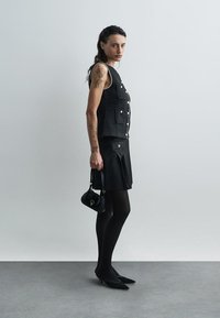 Γυναίκα με τατουάζ στα χέρια, φορώντας μαύρο χωρίς μανίκια γιλέκο, μαύρη φούστα, καλσόν και ψηλές γόβες, κρατώντας μια μικρή μαύρη τσάντα μπροστά σε έναν απλό φόντο.