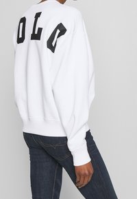 Bílá mikina s černými blokovými písmeny tvořícími nápis „LO“ na zádech. Má volný střih, žebrované manžety a kulatý výstřih.