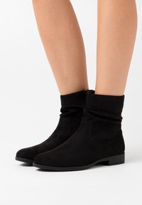 Bottines noires en daim souple. Design ample avec un petit talon carré et une fermeture éclair sur le côté. Bout rond et texture lisse.