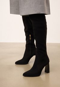 Cizme negre, până la coapsă, cu vârf ascuțit, realizate dintr-un material asemănător suedeului. Acestea au un detaliu cu fermoar auriu și un toc masiv.