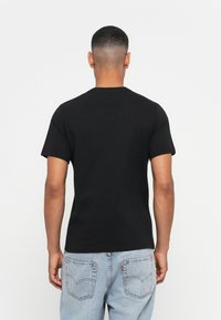 Man with short hair wearing a plain black t-shirt and light blue jeans, standing facing away against a white background.