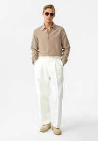 Beige Hemd mit Knöpfen und weiße, gefaltete Hosen. Das Outfit hat eine lockere Passform und wird mit beigen Freizeitschuhen und Sonnenbrille kombiniert.