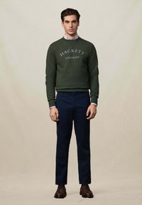 Grüner Sweatshirt mit dem Text "HACKETT LONDON", kombiniert mit dunkelblauen Hosen und braunen Schnürschuhen. Das Model steht mit den Armen an den Seiten.