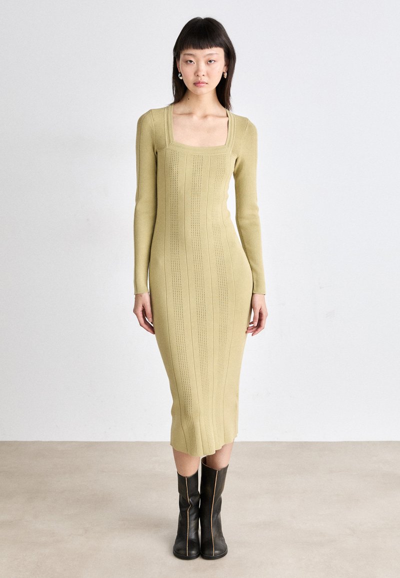 Knit, long-sleeve dress in light olive green with a square neckline, featuring vertical ribbing and a fitted silhouette; paired with knee-high black boots.