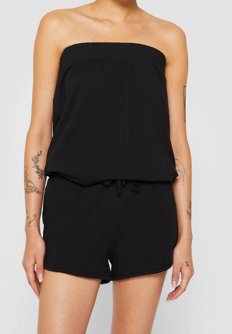 Femme portant une combinaison noire bustier avec un short, montrant des tatouages sur les bras et un teint clair sur un fond gris uni.