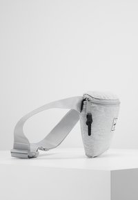 Grå tygcrossbodyväska med texturerad yta, dragkedjestängning och justerbar rem, med en liten logotypmärke på framsidan.