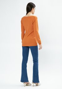 Oranger Pullover mit rundem Ausschnitt, gerippten Bündchen und Saum; kombiniert mit blauen Schlagjeans und hellen High Heels.