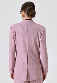 Maßgeschneiderter pinker Blazer aus strukturiertem Stoff, mit Reverskragen und dreiknopfigen Manschetten. Der Rücken weist eine zentrale Naht und einen Schlitz auf.