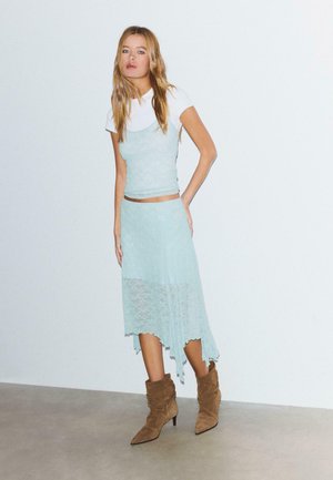 Junge Frau steht, trägt ein hellblaues Spitzenkleid über einem weißen T-Shirt mit braunen Wildleder-Stiefeletten, vor einer schlichten hellen Wand.