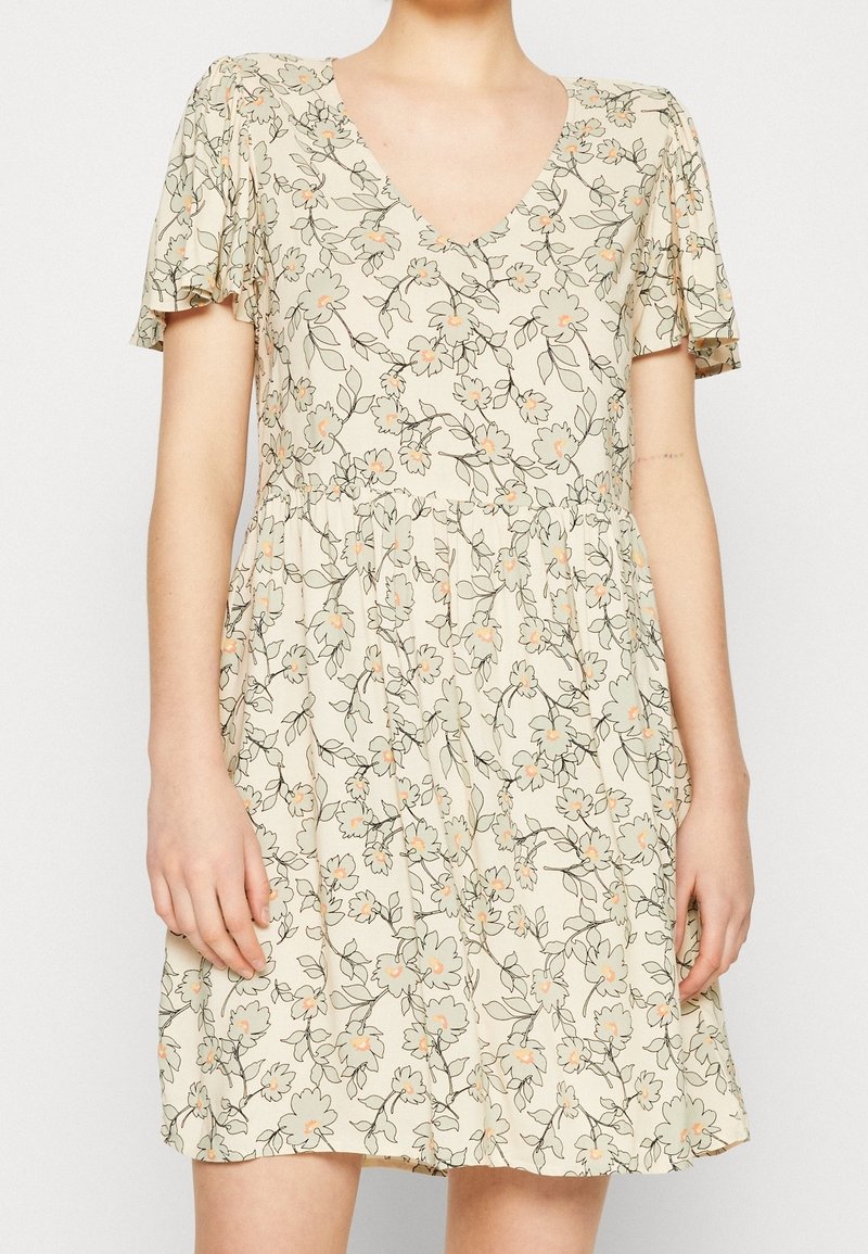 Robe beige à manches courtes avec col en V et petit motif floral en pêche clair et vert, portée par une personne debout montrée du cou jusqu'à mi-cuisse.