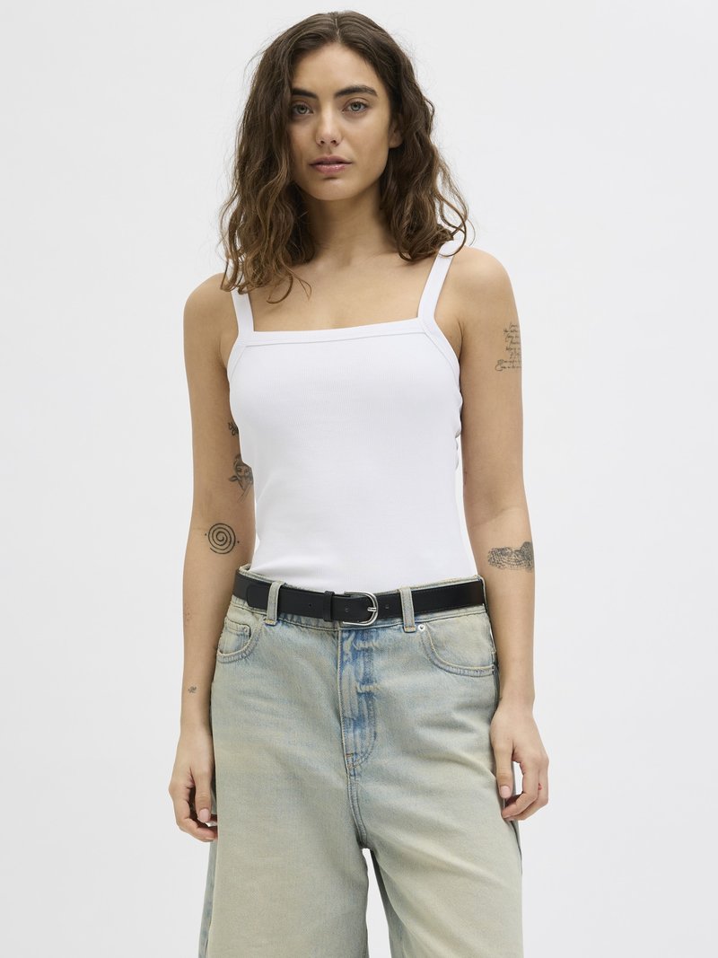 Jeune femme aux cheveux bruns ondulés portant un débardeur blanc à encolure carrée et un jean en denim bleu clair avec une ceinture noire sur un fond uni.