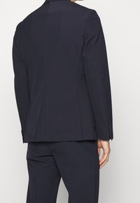 Mörkblå skräddarsydd blazer, strukturerad design, slät textur, en slits bak, knappar med tre knappar vid ärmarna, rena linjer.