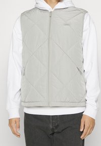 Hellgraue Steppweste mit Rautensteppung, Reißverschluss vorne und Seitentaschen, getragen über einem weißen Hoodie. Glatte Textur.