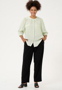 Blusa verde chiaro con bottoni e maniche a palloncino, tessuto testurizzato; abbinata a pantaloni neri larghi e mule nere aperte.