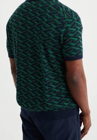 Muž oblečený v košili s krátkým rukávem s geometrickým vzorem v zelené a navy barvě a v navy kalhotách, zobrazený zezadu.