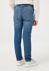 Niebieskie dżinsy o gładkiej fakturze i elastycznym pasku, z dwoma tylnymi kieszeniami i dopasowanym krojem, zestawione z kolorowymi, irydyscentnymi sneakersami.