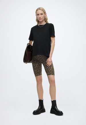 Femme debout portant un T-shirt noir, un short de cyclisme à motif léopard, des bottes noires épaisses, tenant un sac en cuir marron foncé dans sa main gauche.