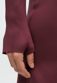 Vinrött figursytt klänningstyg med slät textur; närbild av en hand som håller i ärmen, med en slitsdetalj för tumgrepp.