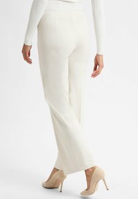 Vrouw in off-white broek en beige hoge hakken, staand met é�én been iets achter het andere gekruist, van achteren gezien.