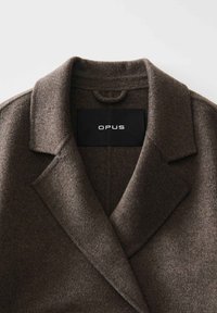 Bruine wollen jas met een geknipte kraag, een enkele knoopsluiting en een zachte textuur, met een "OPUS" label in de nek.