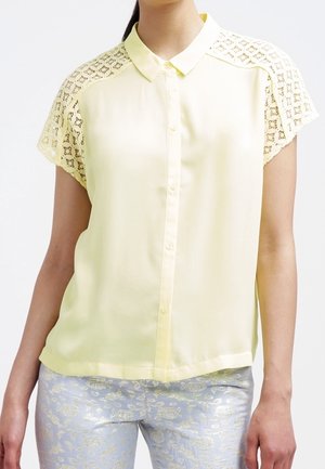 Frau trägt blassgelbe Kurzarmbluse mit Spitzen-Schultern und hellblaue metallisch-florale gemusterte Hose.