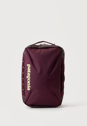 Bolso rectangular Patagonia de color púrpura oscuro con asa superior, logo vertical en el frente y cierre de cremallera sobre un fondo claro y liso.