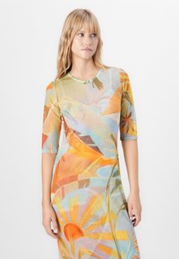 Vrouw met blond haar in een kleurrijke, semi-doorzichtige jurk met abstracte zon- en bladerenpatronen, staand tegen een effen witte achtergrond.