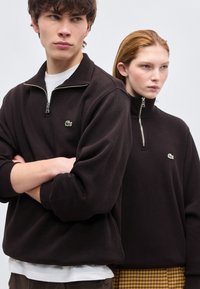 Νεαρός άνδρας και γυναίκα που φορούν σκούρες μπλούζες με φερμουάρ μέχρι το στήθος και πράσινα λογότυπα κροκόδειλου, στέκονται κοντά με ουδέτερες εκφράσεις, ανοιχτό φόντο.