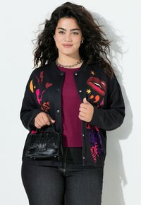 Schwarze Bomberjacke mit lebendigen grafischen Aufdrucken, langen Ärmeln und Druckknopfverschluss. Wird über einem bordeauxroten Oberteil getragen, passend zu dunklen Jeans und einer schwarzen Tasche.