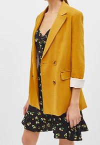 Senapsgul kavaj med dubbelknäppning, snedskurna slag och uppvikta ärmar, lagd över en svart blommig klänning med volanger.