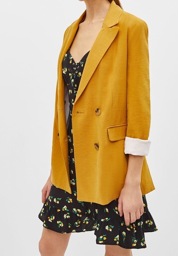 Senapsgul kavaj med dubbelknäppning, snedskurna slag och uppvikta ärmar, lagd över en svart blommig klänning med volanger.