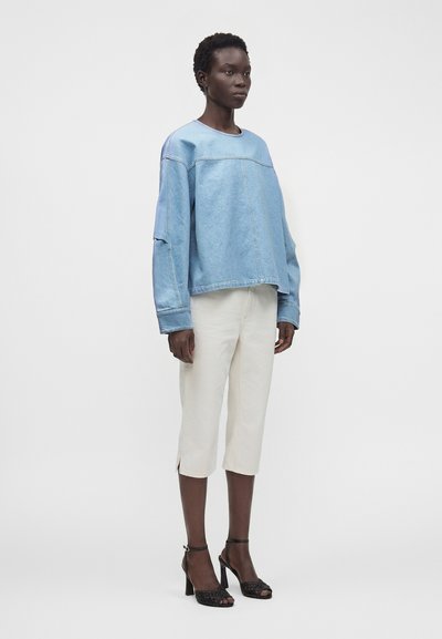 Femme portant un sweat-shirt en jean bleu clair oversize, un pantalon blanc court et des sandales noires à talons hauts, debout sur un fond gris uni.
