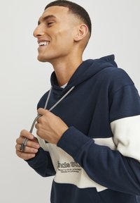Sudadera azul marino con una franja crema, con cordón de ajuste y detalles en plata, y un logo en la parte delantera. Tejido suave y corte relajado.