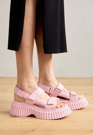 Piernas con pantalones negros cortos y sandalias de plataforma gruesa rosas con correas anchas sobre un suelo de madera clara.