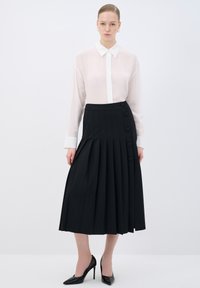 Blusa bianca trasparente con design a bottoni, abbinata a una gonna nera plissettata con bottoni laterali e una texture liscia. Tacchi alti neri.