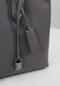 Gros plan d'un coin de sac en cuir gris foncé texturé avec un cadenas métallique noir et une étiquette en cuir embossée du nom de la marque ALDO.
