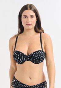 Soutien-gorge noir à pois avec des pois argentés, présentant un design de bonnets structurés, des bretelles ajustables et une bande centrale froncée. Texture de tissu douce.