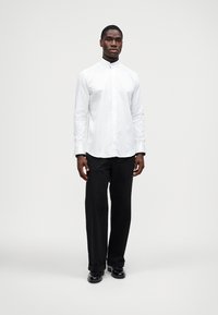 Hvit langermet skjorte med stående krage, sammen med svarte bukser med rette ben og svarte sko, minimalistisk design uten mønstre.