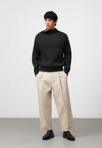 Maglione a coste verde scuro con colletto alto, pantaloni beige plissettati e scarpe nere. Design minimalista, tessuto texturizzato, vestibilità su misura.