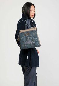 Sac à main bleu avec un design texturé, orné de broderies et d'une fermeture éclair en bronze, porté par une personne dans un pull sombre et texturé.