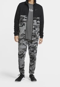 Veste zippée noire avec des accents camouflage gris, associée à un T-shirt gris et un pantalon de jogging camouflage assorti. Des chaussures de sport noires complètent la tenue.