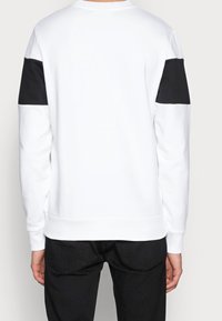 Felpa bianca con dettagli neri sulle spalle, polsini e orlo a costine, e una finitura morbida e texturizzata. Vista posteriore mostrata.