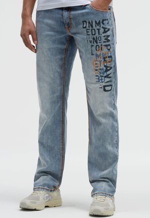 Man die lichtblauwe jeans met donker bedrukte tekst en een wit T-shirt draagt, staande met één hand in de zak en beige sneakers.