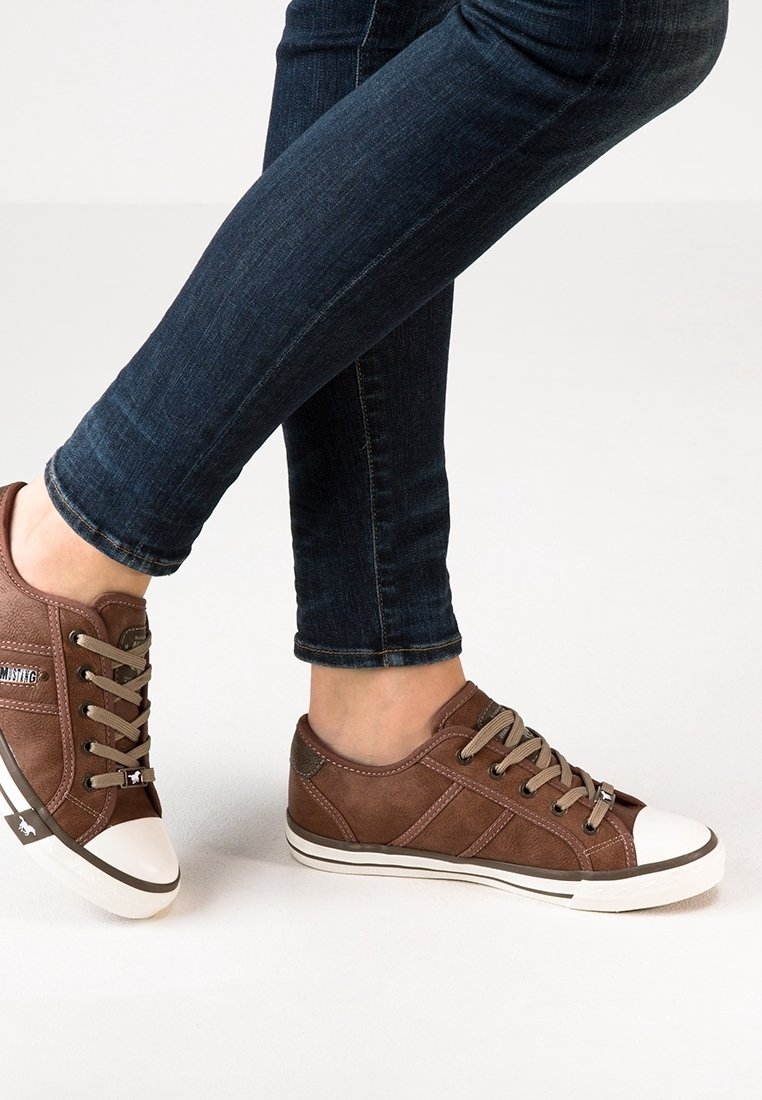 Personne portant un jean bleu foncé et des baskets basses marron à bouts blancs, debout sur un sol clair, jambes croisées.