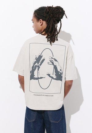 Homme aux dreadlocks de dos portant un t-shirt beige avec des mains graphiques s'étendant vers un ovale et un texte en bas.