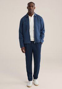 Jūras bruņu krāsas sukta jaka ar divām priekšējām kabatām, kombinēta ar jūras bruņu krāsas biksēm un baltām sporta kurpēm. Gluda tekstūra, ikdienišķs dizains un ērtas piegriezums.